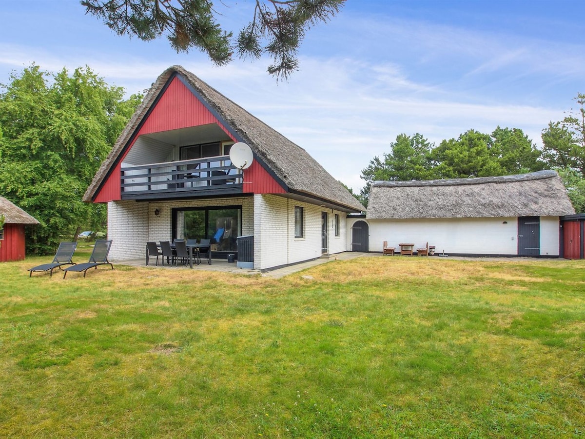 A spacious two-story house features a thatched roof and a covered balcony, surrounded by a well-maintained lawn. Outdoor seating areas are available, including lounge chairs and a dining set. Nearby, a separate structure with red accents complements the tranquil natural setting.