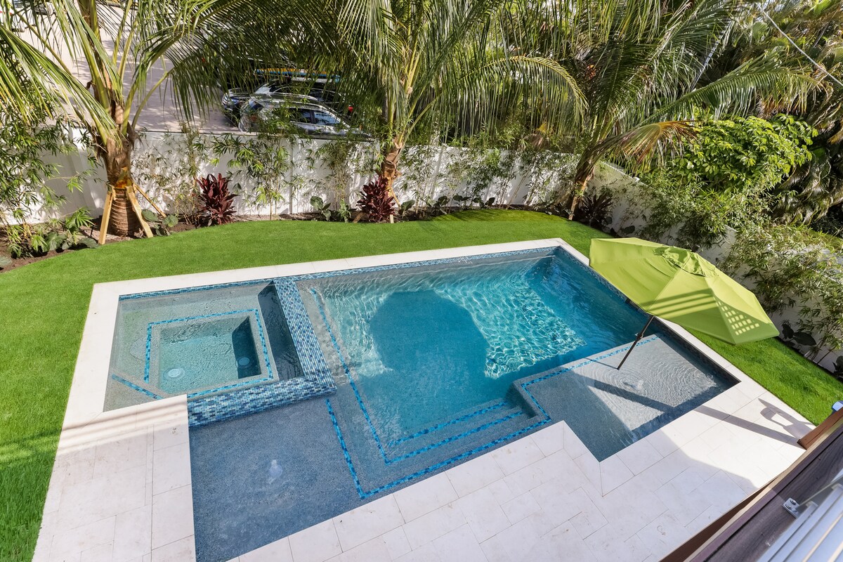 A serene outdoor space features a heated pool with an attached spa, surrounded by lush greenery and palm trees. A sun shelf with a vibrant green umbrella is positioned near the pool. The area is highlighted by a well-maintained lawn and patio tiles.