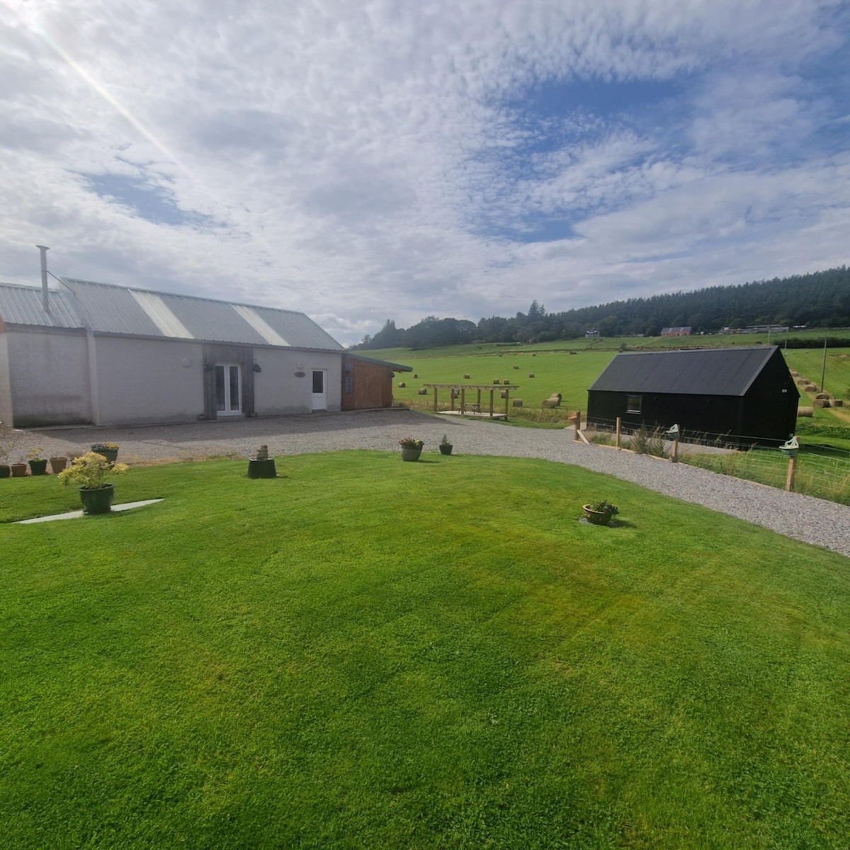 The exterior of the cottage is presented, showcasing a well-maintained grassy area with potted plants. A gravel path is visible leading to the building. In the background, a shed can be seen along with lush green fields and a clear sky, enhancing the rural scenery.