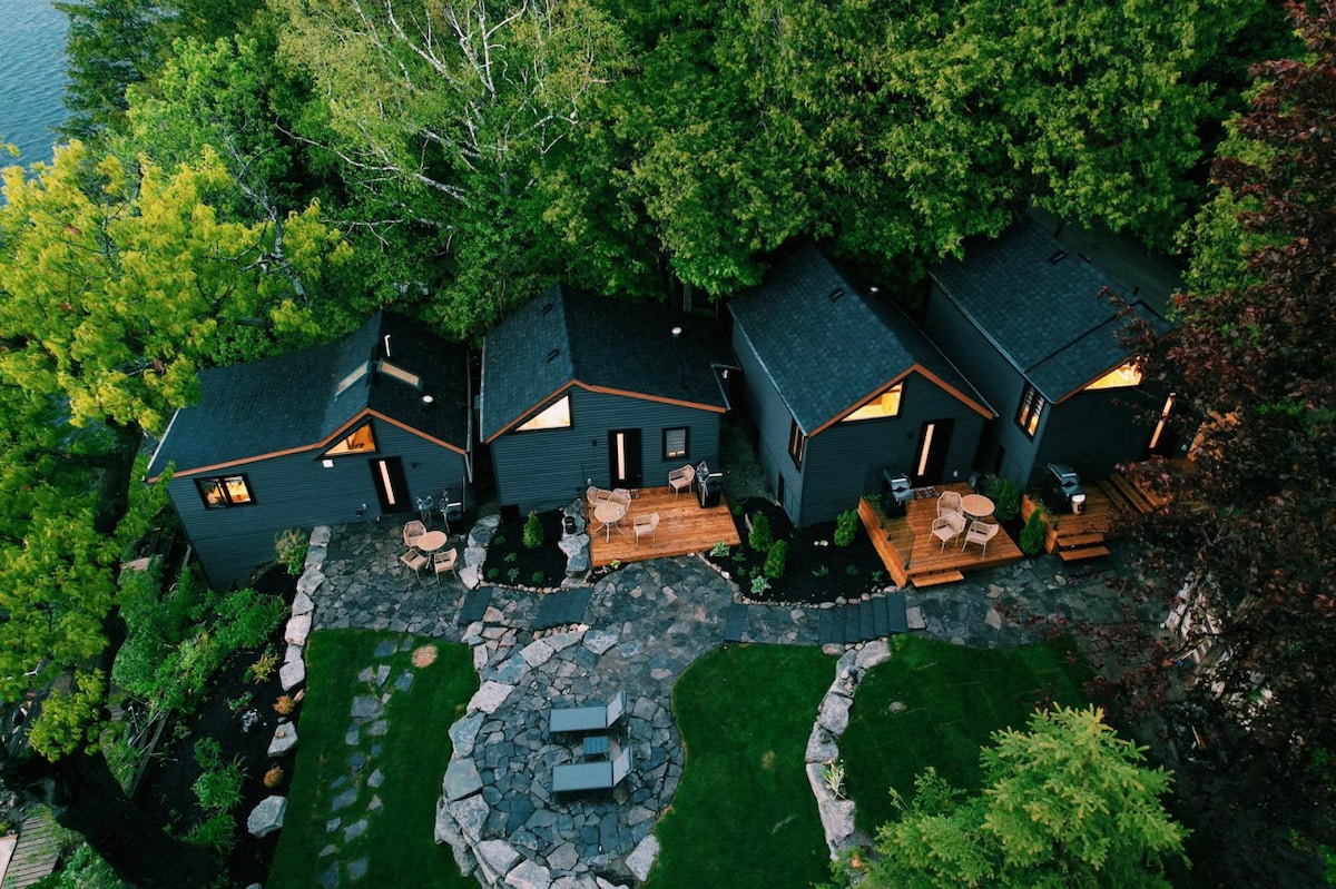 An aerial view showcases three modern cabins nestled among lush greenery, creating a harmonious blend with nature. Stone pathways meander through a landscaped yard featuring manicured grass and seating areas. The cabins' decks are equipped with outdoor chairs, inviting relaxation by the lake.