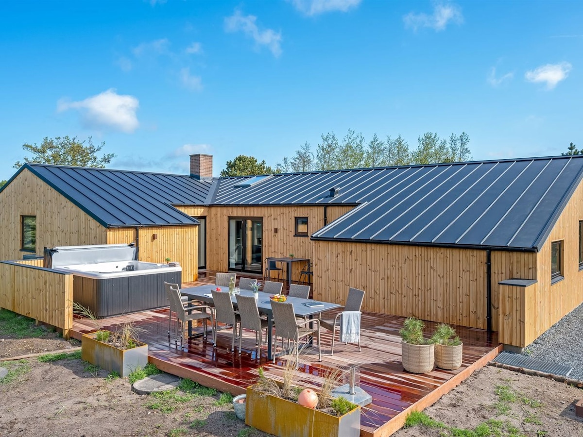 An outdoor seating area is presented with a large wooden table surrounded by chairs, set upon a smooth deck. A hot tub is positioned to the left, adjacent to a single-story wooden structure under a black roof. Greenery complements the natural surroundings.
