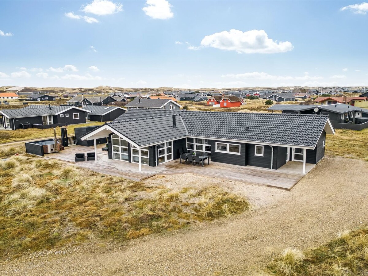An aerial view of a modern single-family house with a dark exterior, surrounded by natural grassy land. A spacious wooden deck features outdoor seating, providing a comfortable area for relaxation. Nearby properties are visible, showing a mix of colors and designs, with mountains in the background.