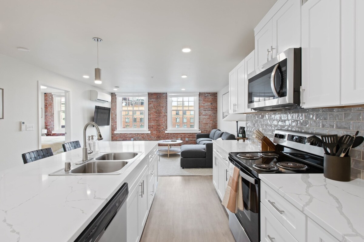 The modern kitchen features white cabinetry and a spacious countertop with a double basin sink. Stainless steel appliances, including a microwave and stove, are visible. Natural light fills the area from large windows, illuminating the adjacent living space furnished with a sectional sofa.