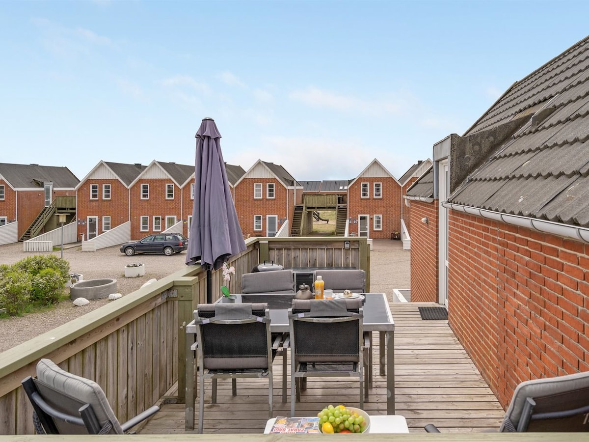 A deck area is presented, featuring a table with chairs, suitable for outdoor dining. A large umbrella provides shade. In the background, several red brick houses are visible, arranged in a row, with a clear sky above.