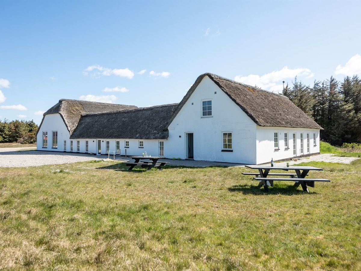The exterior of the charming single-family house is shown, featuring a thatched roof and white-painted walls. A spacious lawn surrounds the property, with picnic tables visible in the foreground. Tall trees line the backdrop, under a bright blue sky with a few clouds.