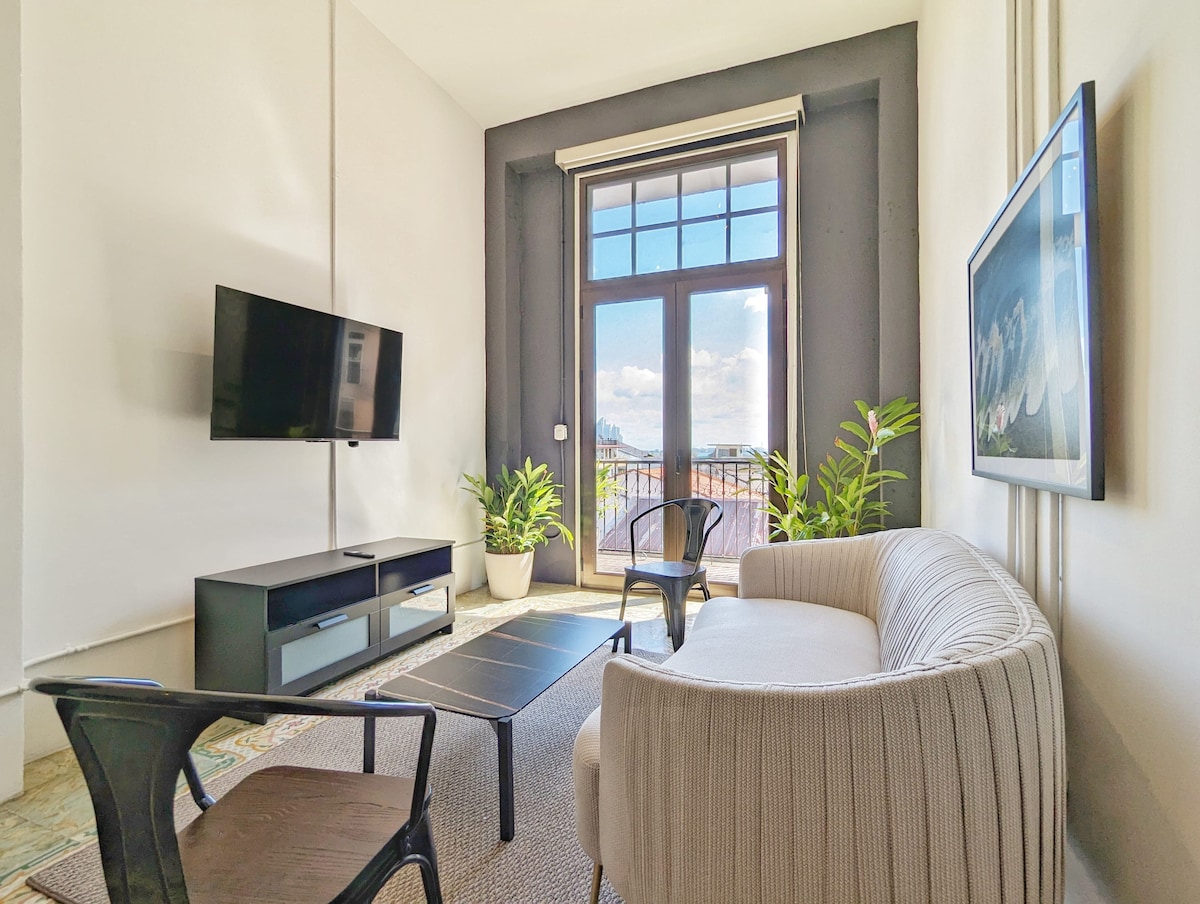 A spacious living area is highlighted by large windows allowing natural light to fill the room. A comfortable sofa faces a sleek coffee table and a wall-mounted television. Decorative plants add a touch of greenery, enhancing the overall ambiance.