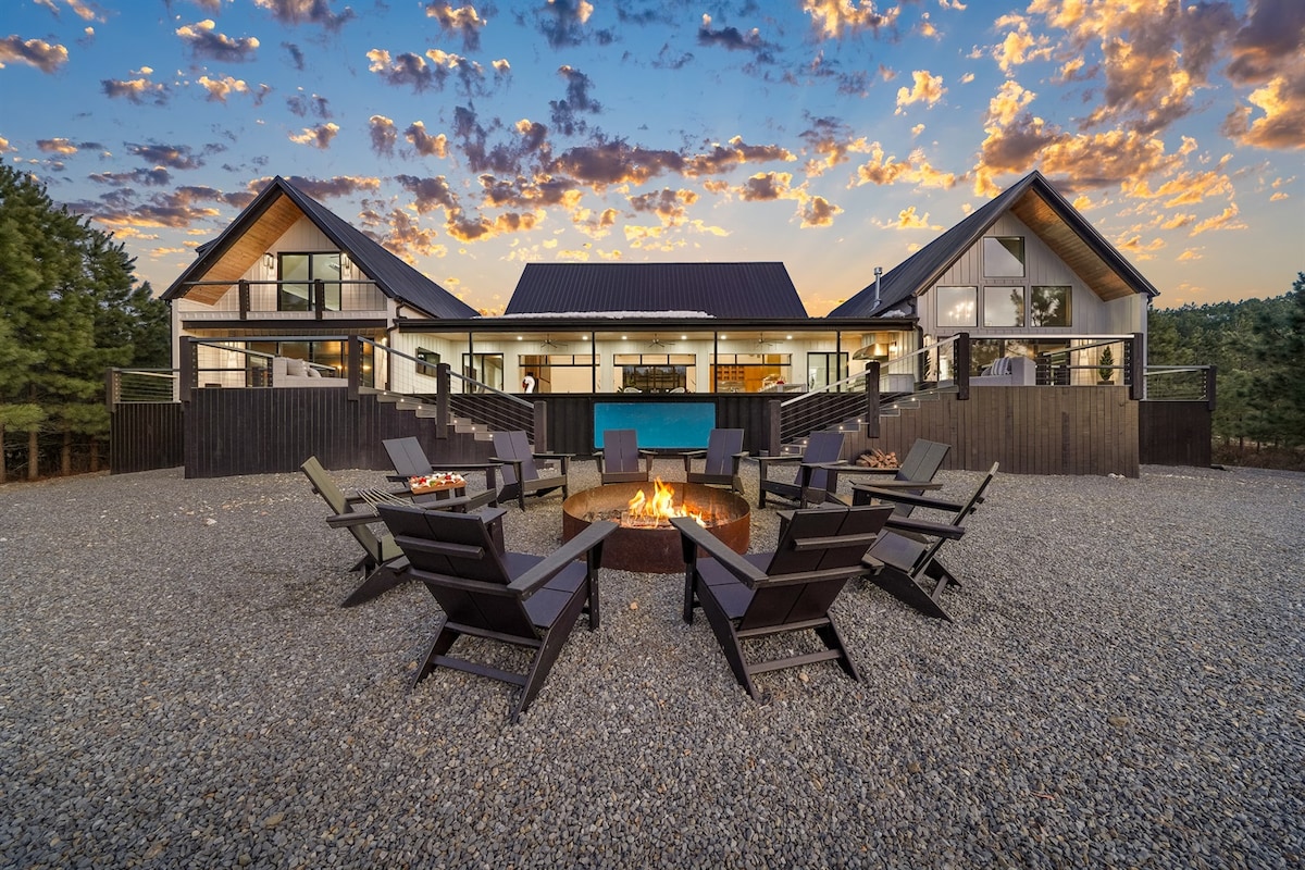 An inviting fire pit area is surrounded by multiple chairs, set against a backdrop of a spacious villa. The villa features expansive windows and a large terrace, with a pool visible, all bathed in warm evening light beneath a colorful sky.
