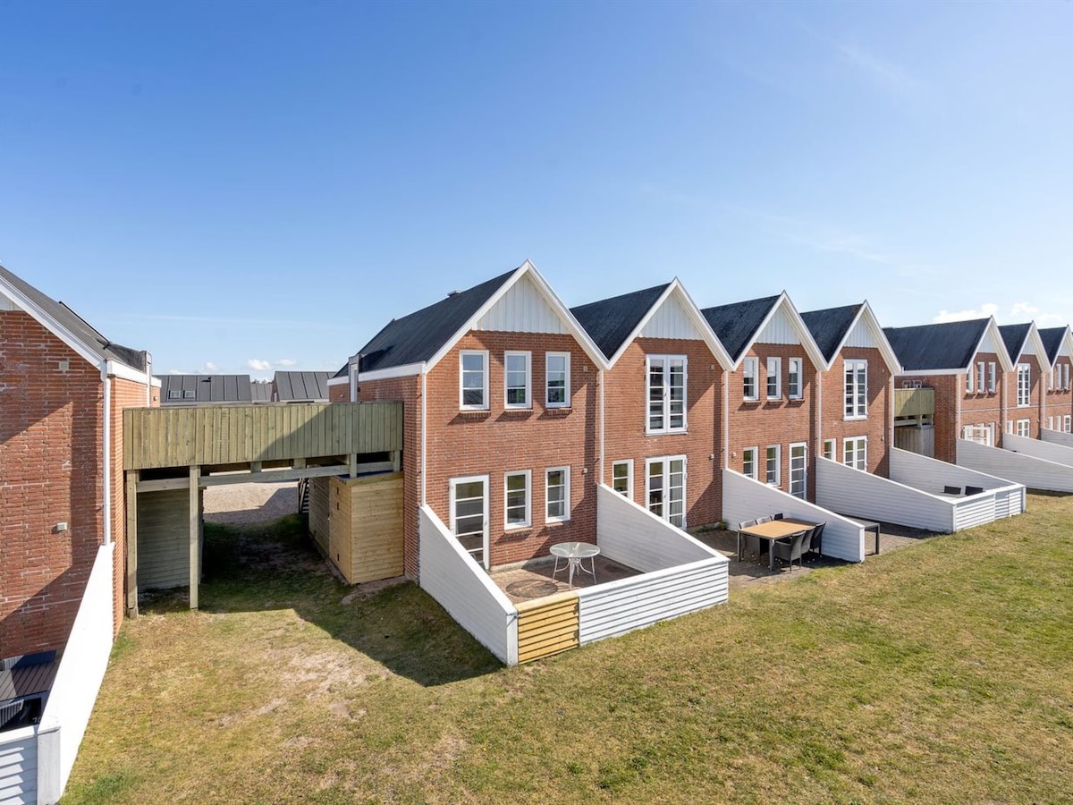 A row of two-story buildings is displayed, featuring modern architecture with sloped roofs and red brick exteriors. Each unit includes large windows and a private outdoor space with seating areas. Green grass surrounds the structures, contributing to a relaxed environment.