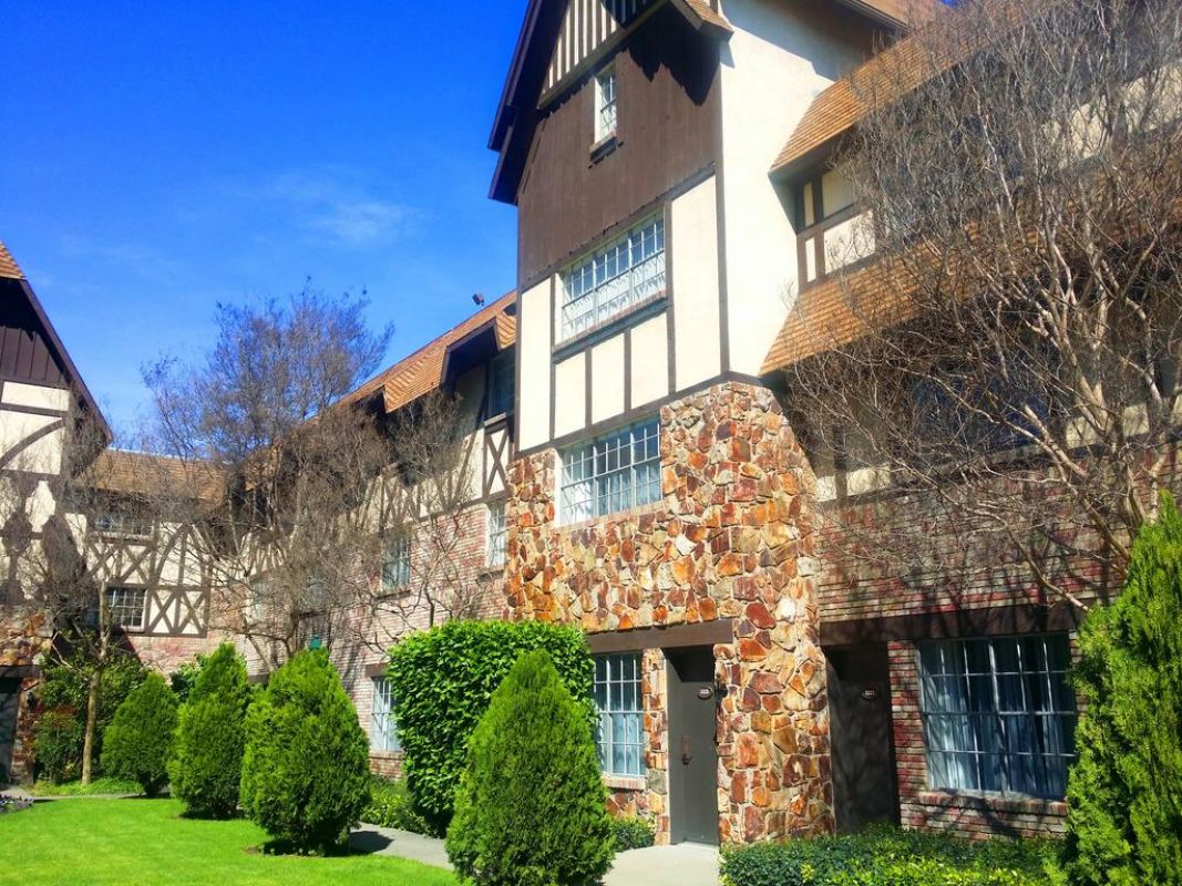 The exterior of the building showcases a charming castle-inspired design with a combination of stone and timber features. Lush greenery surrounds the entrance, with neatly trimmed hedges and vibrant landscaping under a clear blue sky.