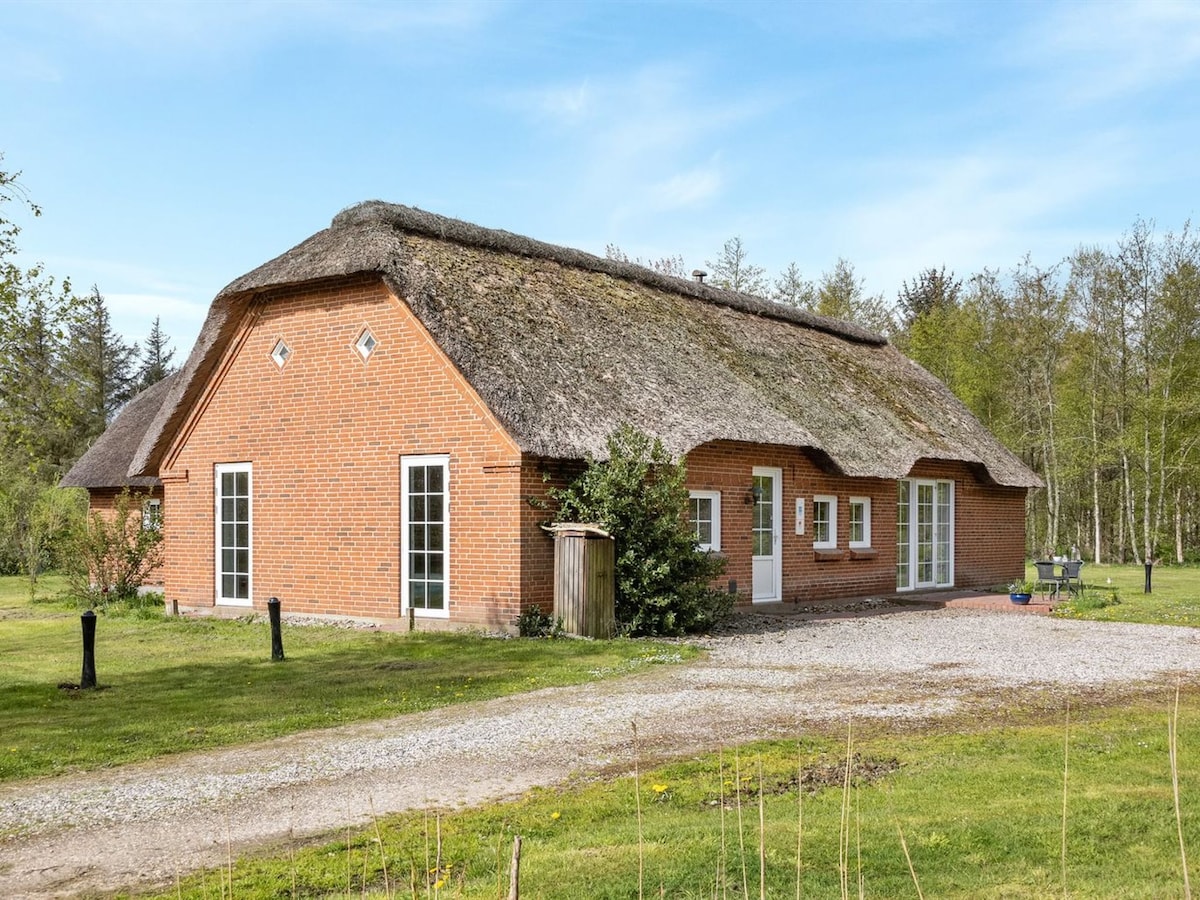A single-family house made of red brick features a traditional thatched roof. Large windows provide natural light, and a small pathway leads to the entrance. Lush greenery surrounds the property, enhancing the tranquil outdoor setting.