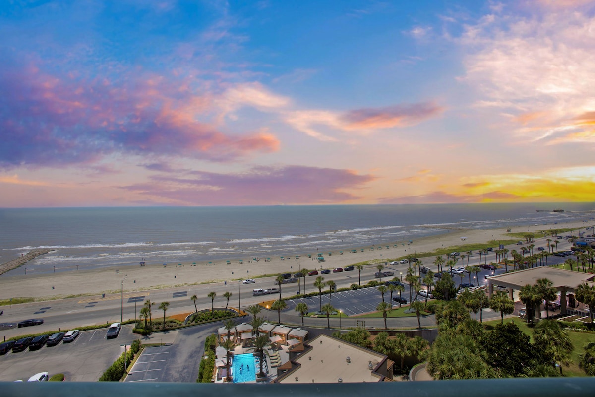 A sweeping coastal view depicts the sandy beach and ocean under a colorful sunset sky. Palm trees line the foreground, while vehicles dot the road alongside the shore. The scene captures a tranquil seaside atmosphere with inviting blue waters and gentle waves.