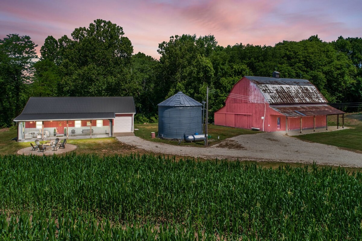 A country property features a red barn and a silos beside a gravel path. Outdoor seating with chairs and a table is positioned near the main residence, surrounded by lush greenery and cornfields. The sunset casts a warm glow over the landscape.