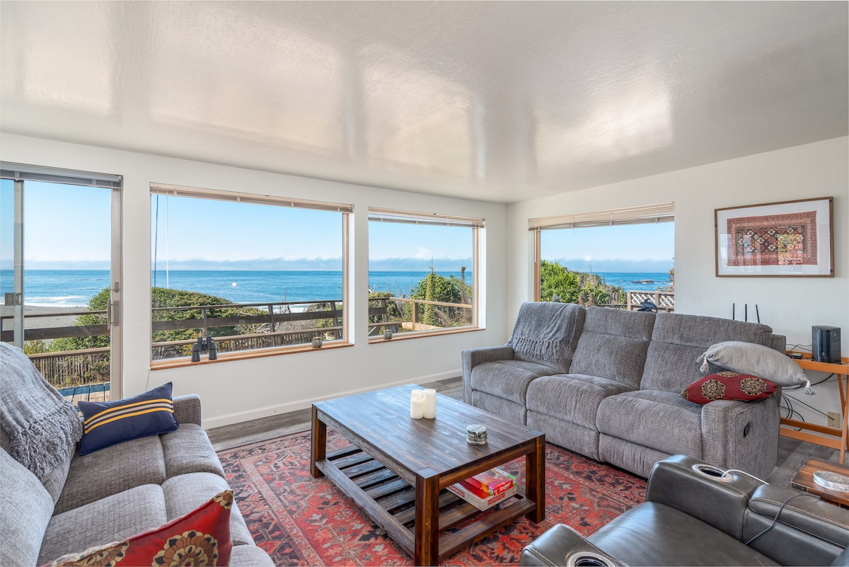 A cozy living room showcases large windows offering sweeping ocean views. Comfortable grey sofas are arranged around a wooden coffee table, while natural light fills the space. A colorful area rug adds warmth, and a nearby console holds entertainment equipment.