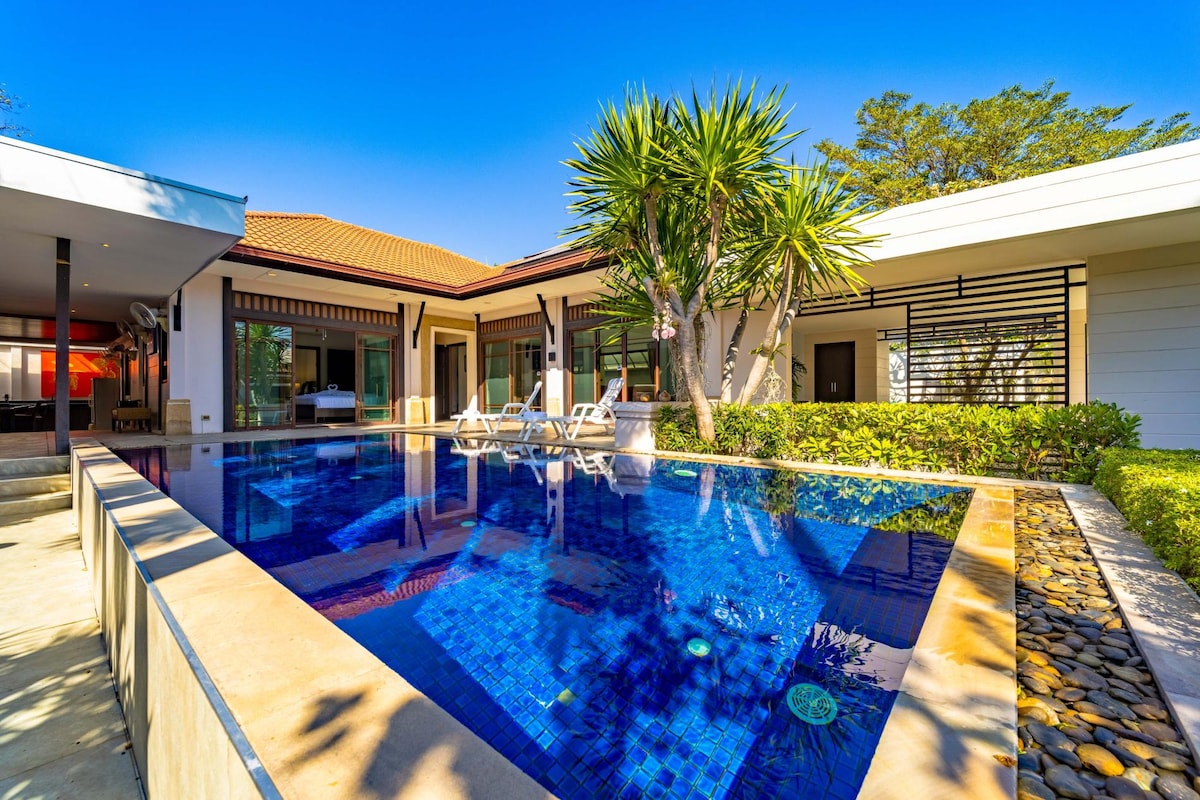 A tranquil pool area is showcased, featuring a clear blue swimming pool bordered by a stone patio. Sun loungers are positioned near the water, while lush greenery and palm trees create a serene environment. The modern villa structure is visible in the background.
