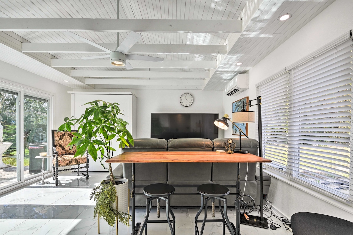 A modern living area features a light wood bar table accompanied by two stools. A gray sofa is positioned facing a wall-mounted television, with natural light flooding through large windows. Indoor plants and a floor lamp add to the inviting space, complemented by a ceiling fan.