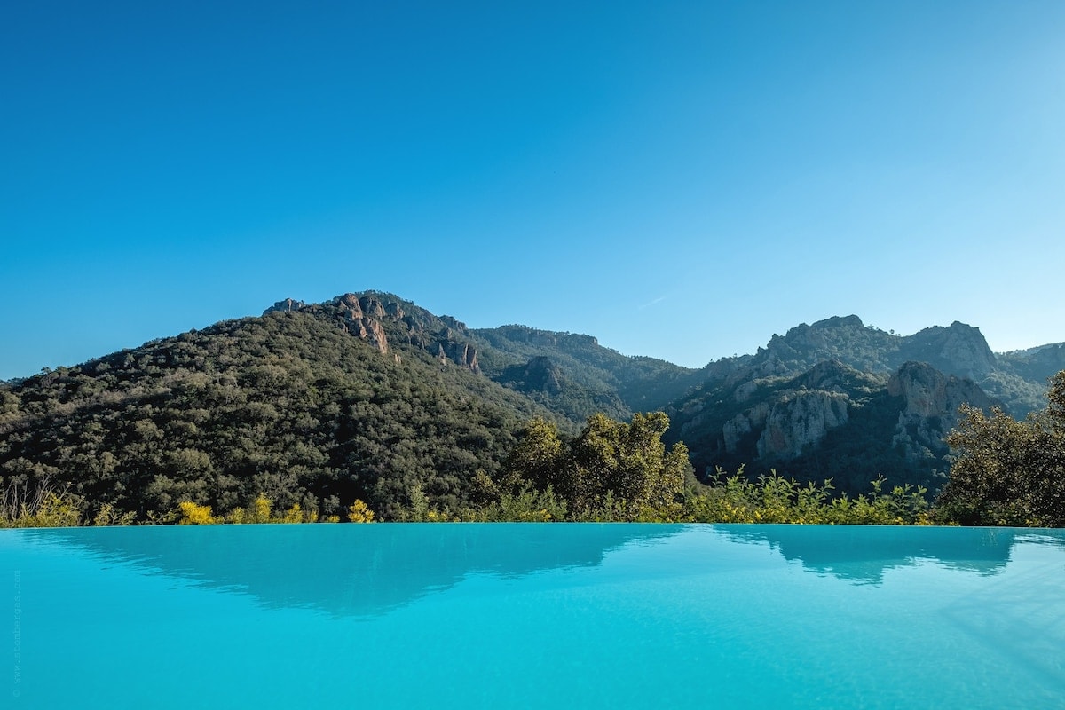 An infinity pool stretches out towards a breathtaking view of rolling hills and rugged mountains. The clear blue water reflects the sky, merging seamlessly with the natural landscape. Lush greenery frames the scene, enhancing the sense of tranquility and connection to nature.