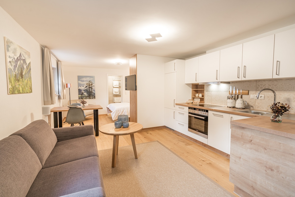A modern living area features a cozy gray sofa and a wooden table with an ergonomic chair. A compact kitchen with white cabinetry and an oven is visible. Natural light brightens the space, enhancing the warm wooden flooring and decor pieces that reflect the surrounding natural beauty.
