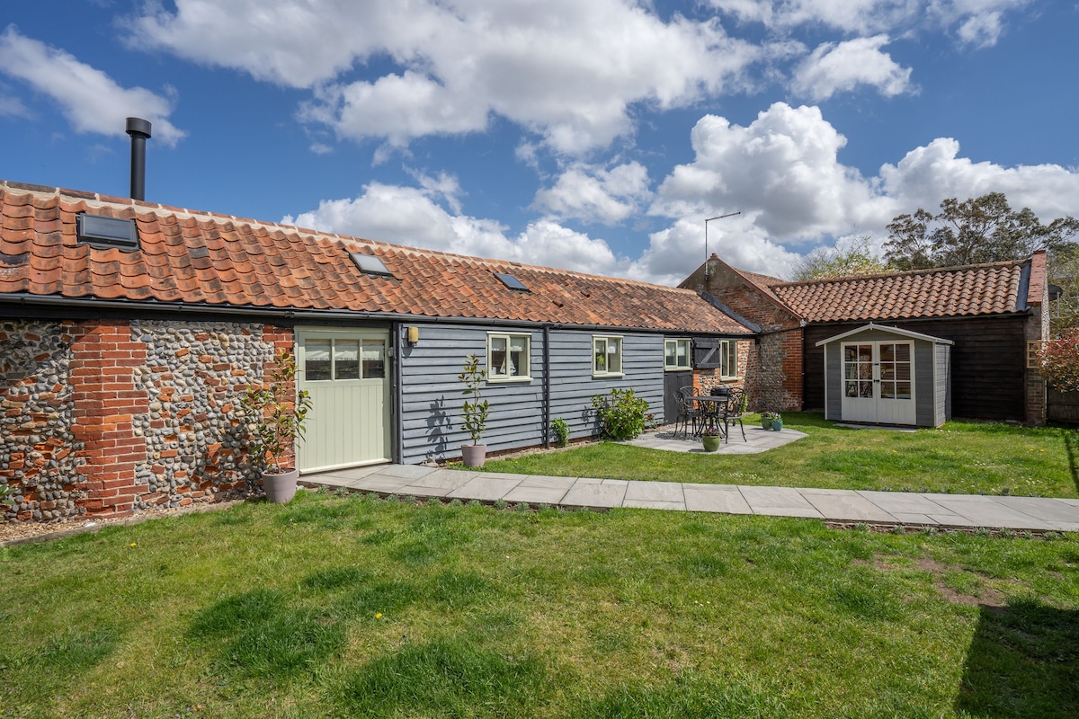 The exterior of Natterjack is showcased with rustic stone and wood siding. A pathway leads to the front door, surrounded by a grassy area. A small patio with seating is visible, framed by blooming plants under a bright sky with puffy clouds.