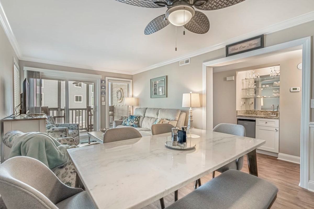 An open family room and dining area is visible, featuring a marble dining table with soft gray chairs. A comfortable sectional sofa and TV are arranged for relaxation. Sliding glass doors lead to a balcony, allowing natural light to enhance the space.