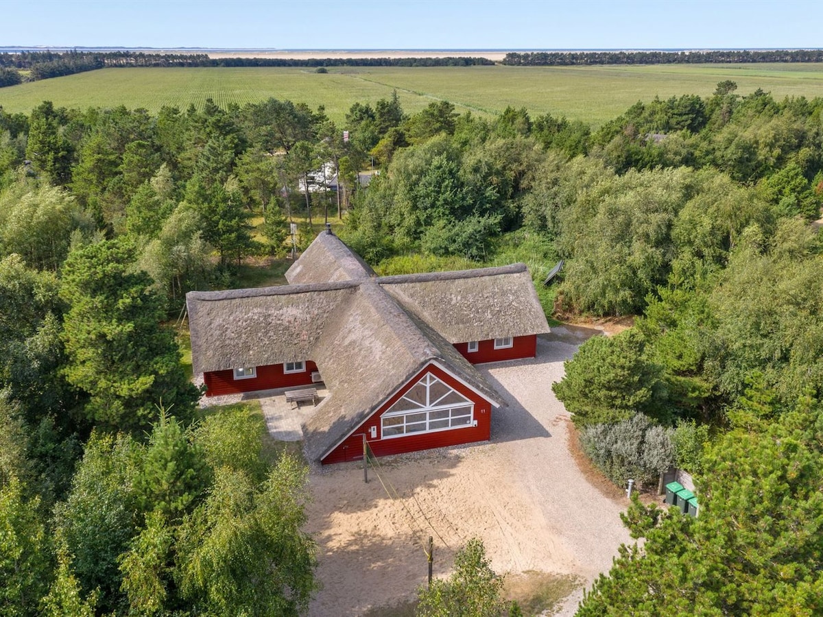 A spacious, single-family house features a thatched roof and red exterior, set amidst lush greenery. The property is surrounded by a natural landscape, with open fields visible in the distance. A gravel drive leads to the entrance, framed by trees and shrubs.