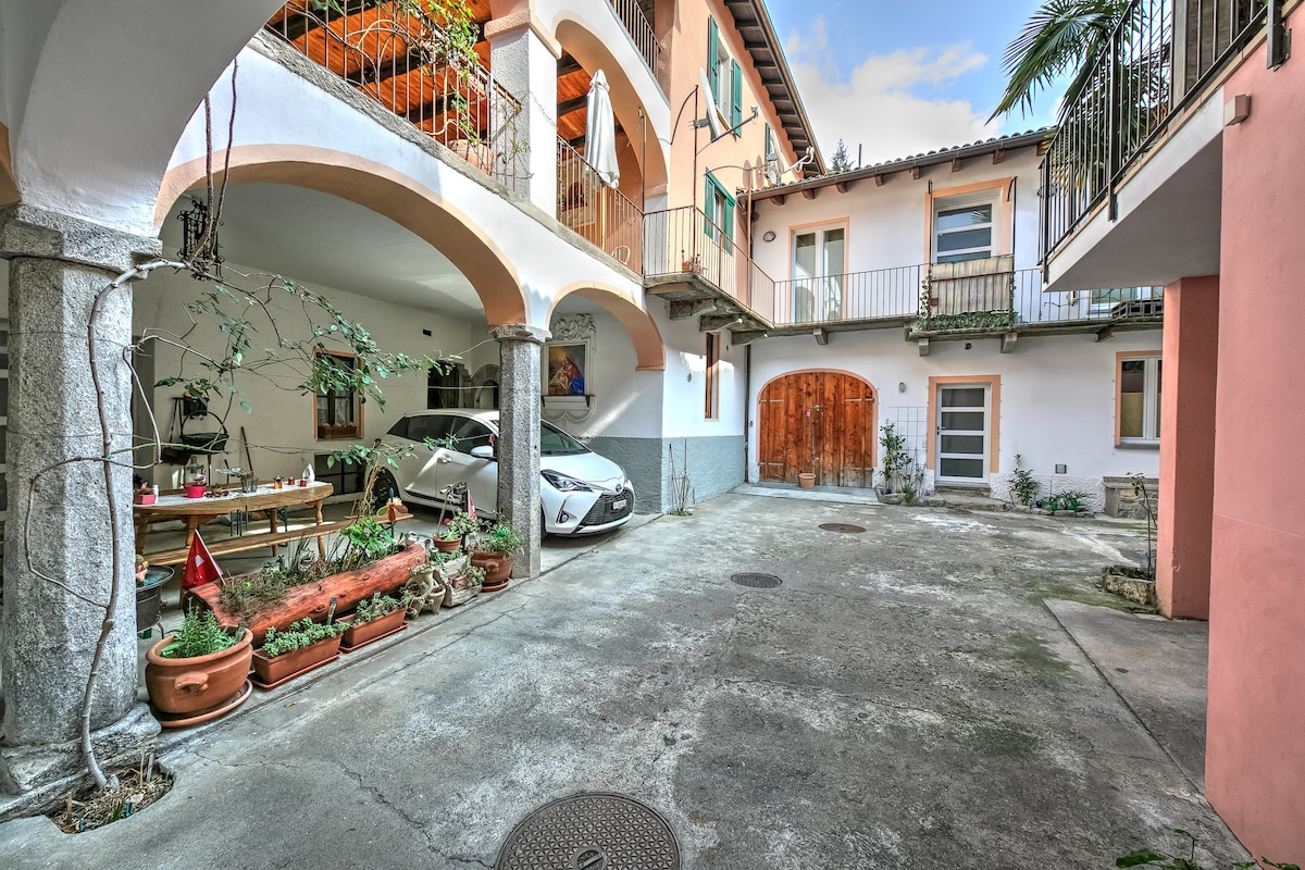A shared courtyard is flanked by two buildings featuring arched doorways and balconies. Potted plants add greenery, while a parked car is visible near a wooden door. Tables and chairs suggest a space for outdoor dining or relaxation.