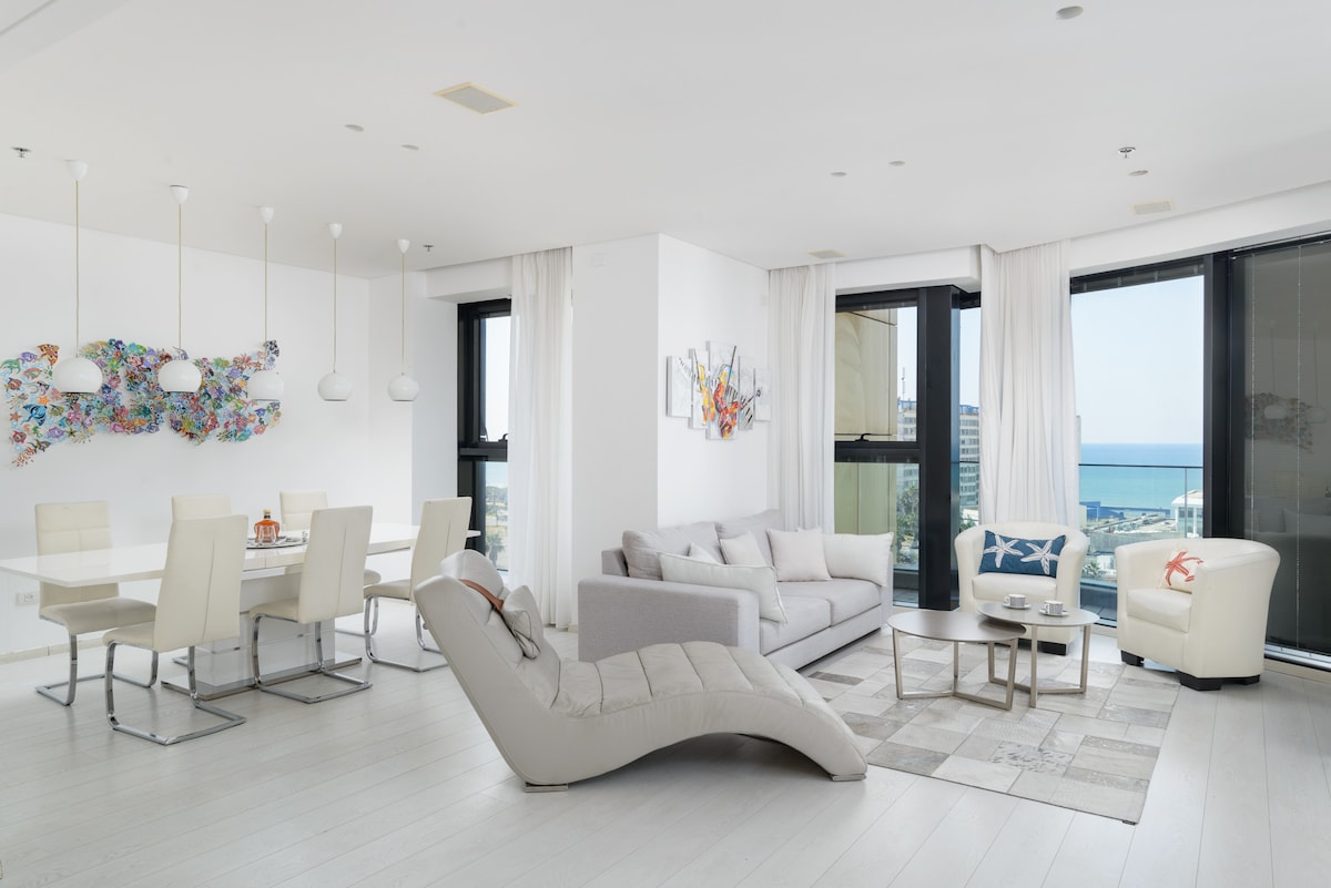 A spacious living area is highlighted by large glass windows offering a sea view. The room features elegant furnishings, including a comfortable light-colored sofa, a lounge chair, and a modern dining table with six chairs. Bright artwork adds color to the white walls.