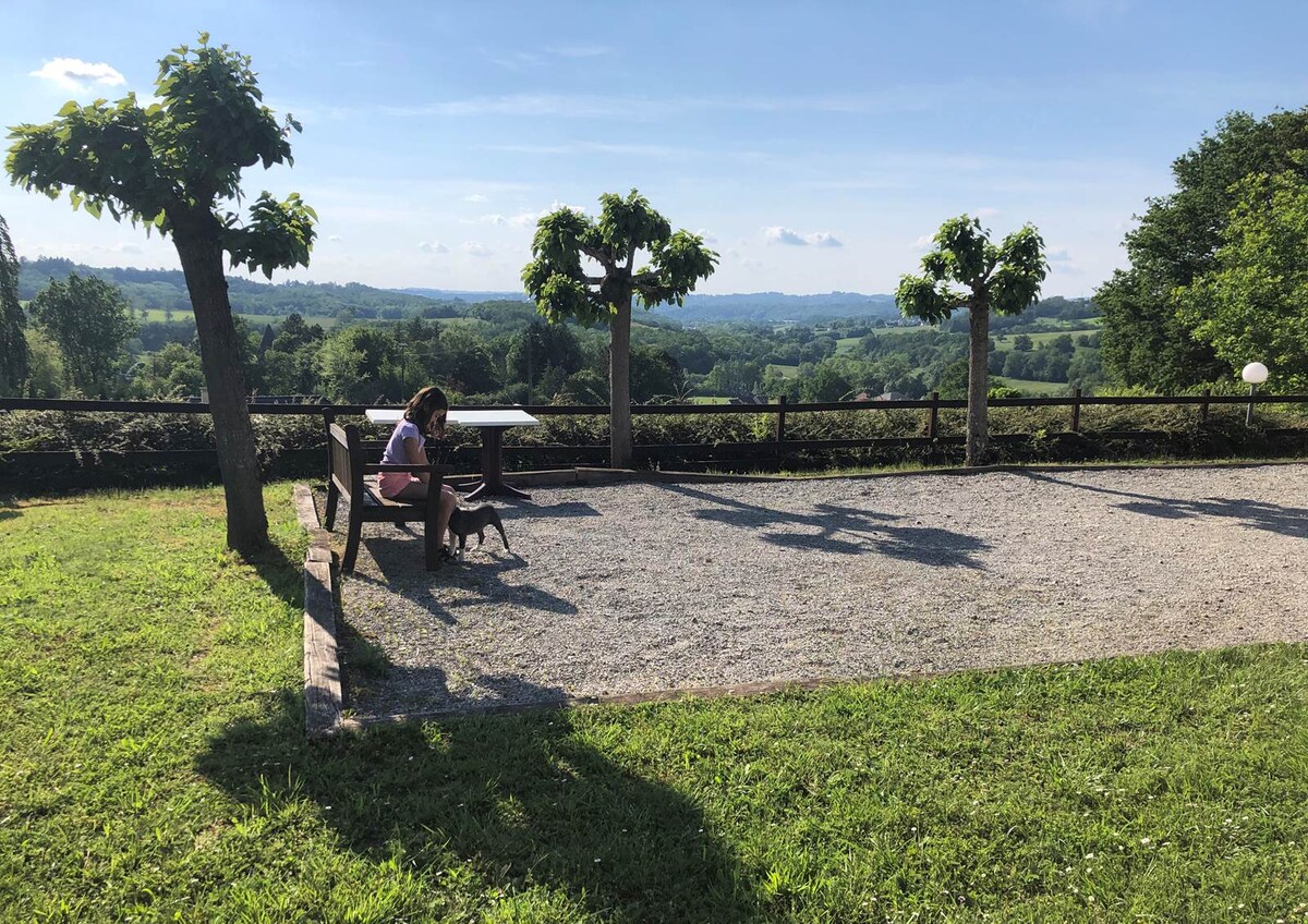 A gravel area is framed by two small trees, with a person seated on a bench alongside a dog. The expansive view of rolling hills and lush greenery stretches into the background, under a clear blue sky with scattered clouds.