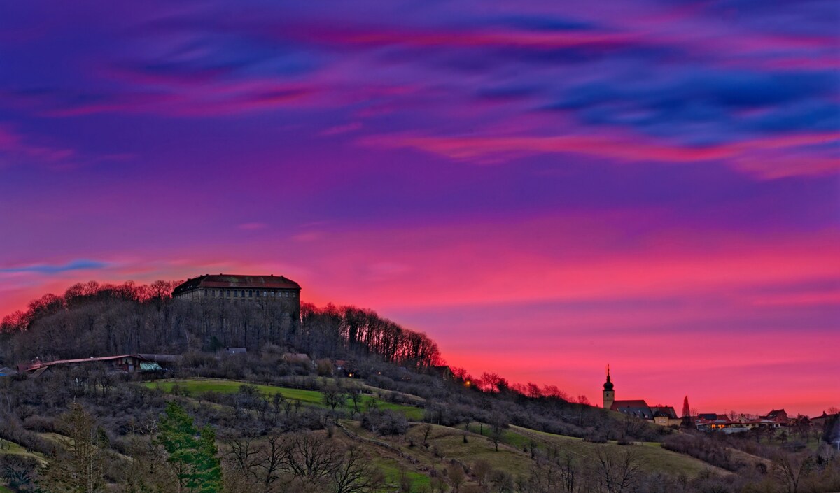 A vibrant sunset paints the sky with shades of pink and purple, providing a stunning backdrop to a hillside. Silhouettes of trees and a historical building are visible, along with a steeple emerging above the landscape, creating a serene ambiance.