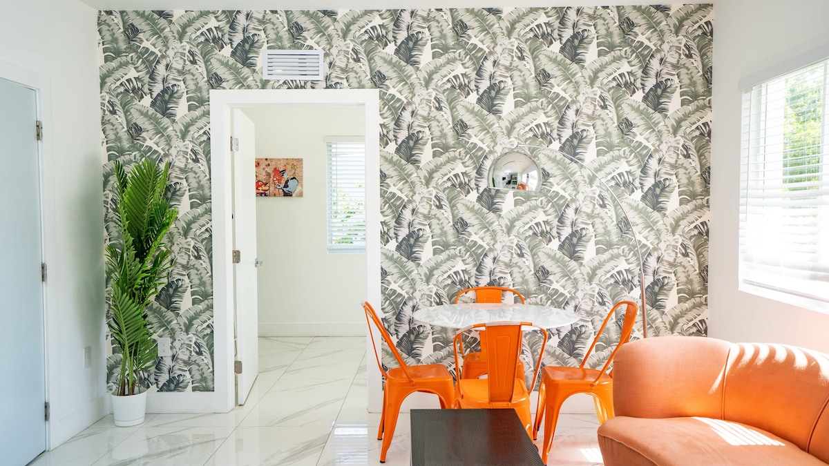 A sunlit lounge area is highlighted by a decorative wall featuring tropical foliage. A marble table is surrounded by four modern orange chairs. A cozy orange sofa is positioned nearby, complemented by natural light streaming through the window adorned with white blinds.