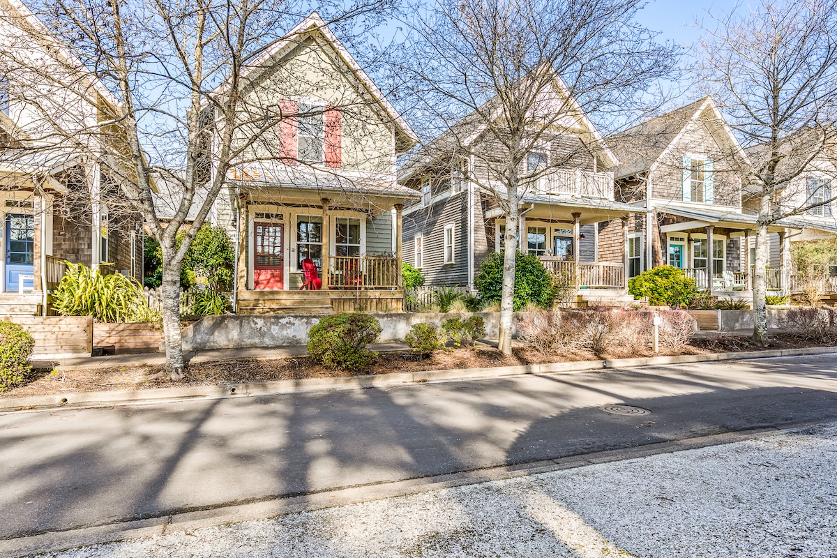 A row of charming coastal homes featuring porches with rocking chairs and well-kept landscaping. Sunlight casts gentle shadows on the pavement, highlighting the serene streetscape. The surroundings are characterized by trees and hedges, contributing to a peaceful neighborhood atmosphere.