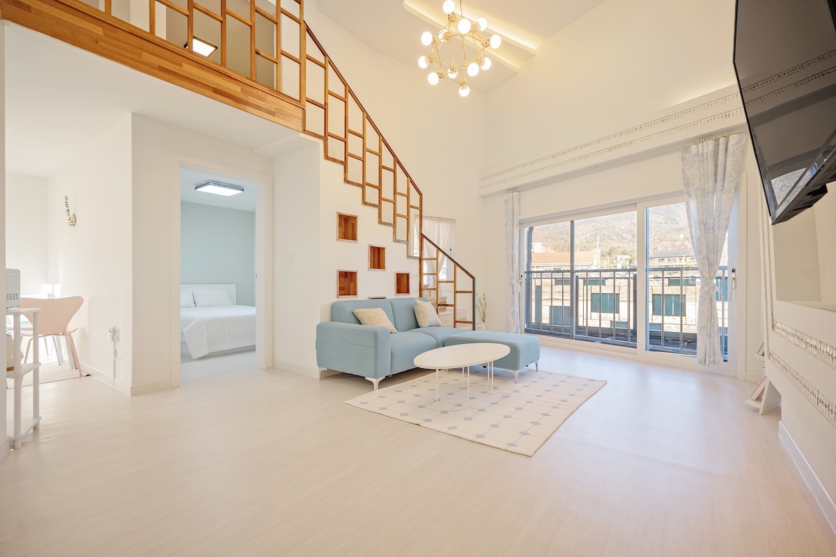 A bright and airy living space features a light blue couch and a circular coffee table. Large windows offer scenic views, while a wooden staircase leads to the upper level. A soft rug adds warmth to the white floor, creating an inviting atmosphere.
