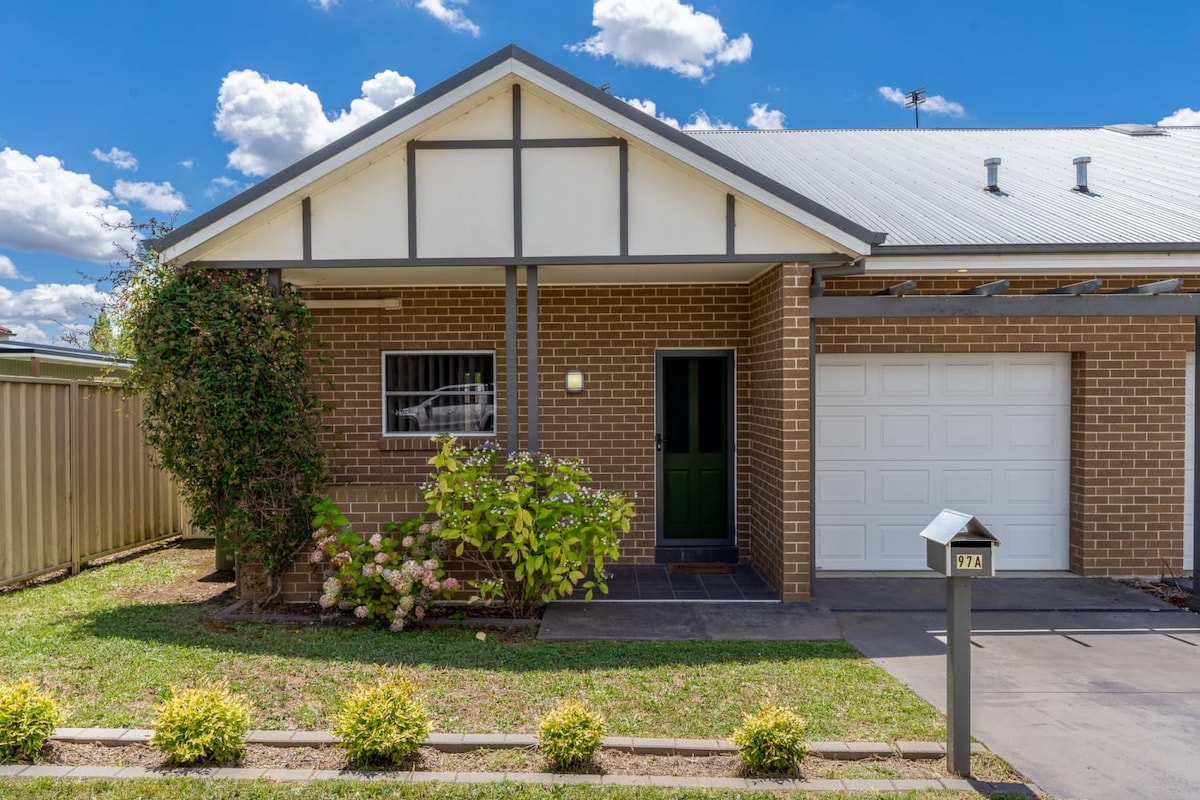 The exterior of the townhouse features a brick façade with a pitched roof. A green front door is framed by a small garden with blooming plants. The attached garage has a single door, and the pathway leads to a well-maintained lawn.