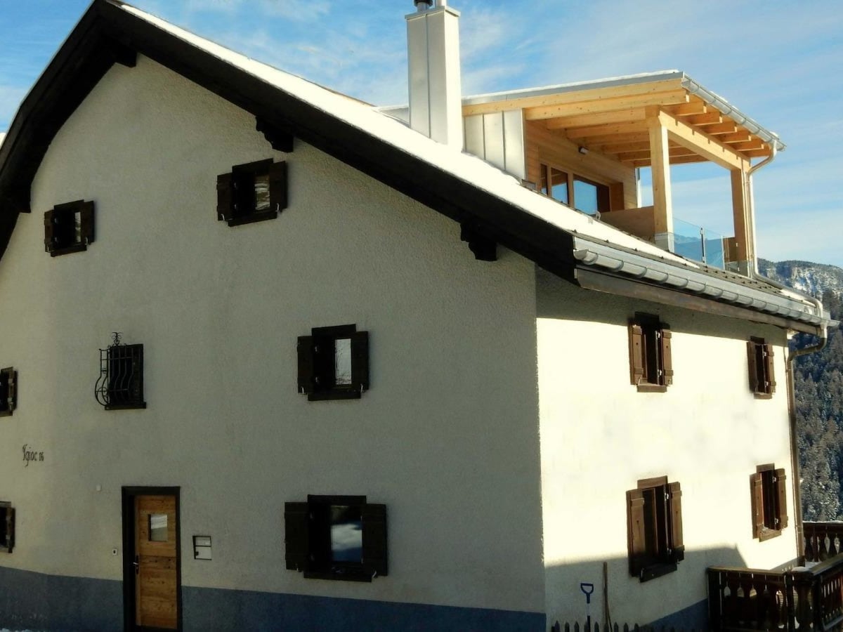 The exterior of the holiday home is shown, highlighting a sloped roof with wooden elements and multiple dark window shutters. A newly built roof terrace is visible at the top, offering a view over the surrounding nature.