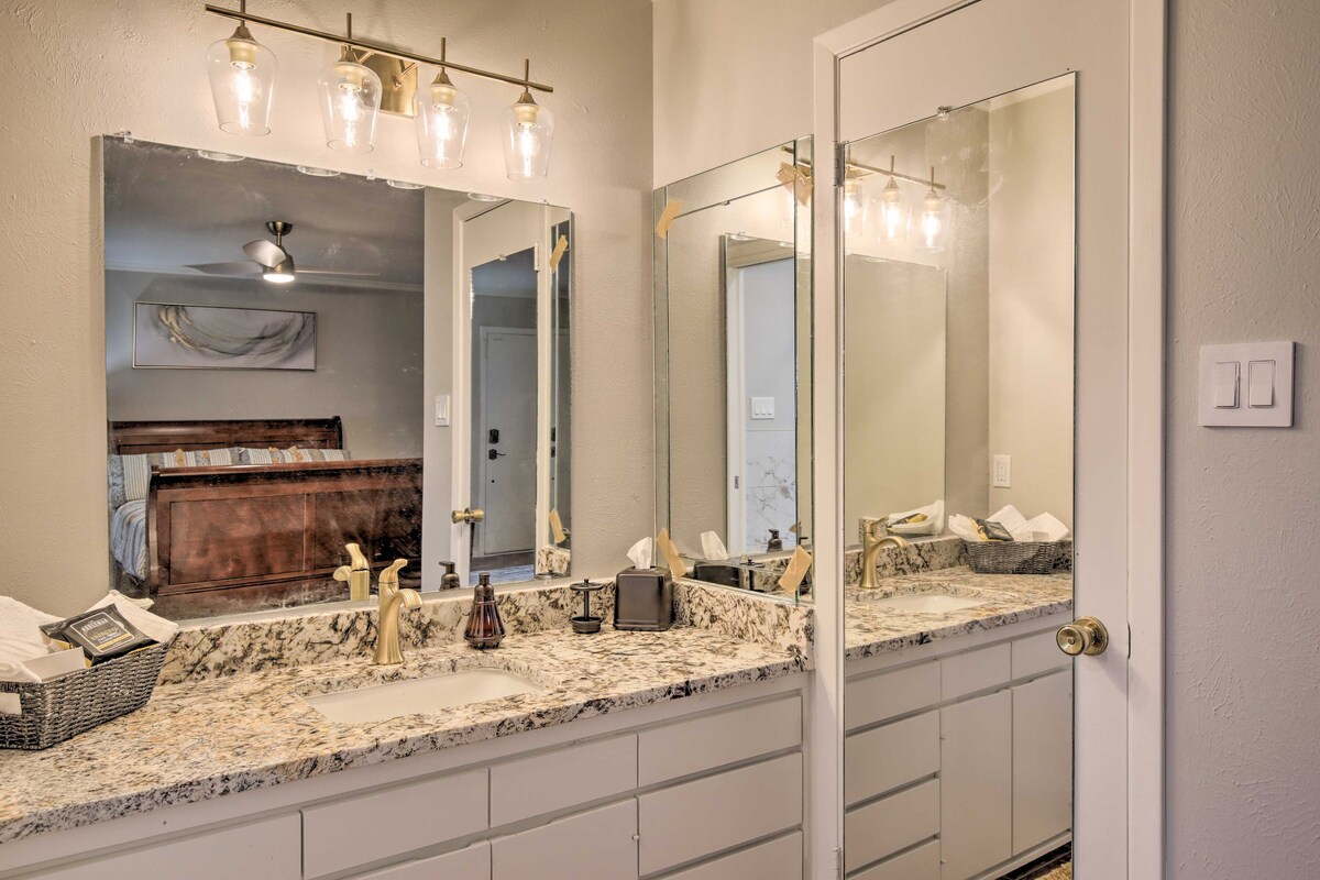 A well-lit bathroom features dual vanities with a granite countertop and ample storage beneath. A large mirror reflects soft lighting from above, while a glimpse of the bedroom can be seen in the background, enhancing the open layout.