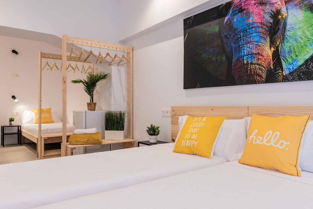 A bright sleeping area is furnished with two spacious beds, each featuring soft white linens and decorative yellow pillows. A colorful elephant artwork decorates the wall, while a small plant adds a touch of greenery. A clothing rack is positioned adjacent to the beds.