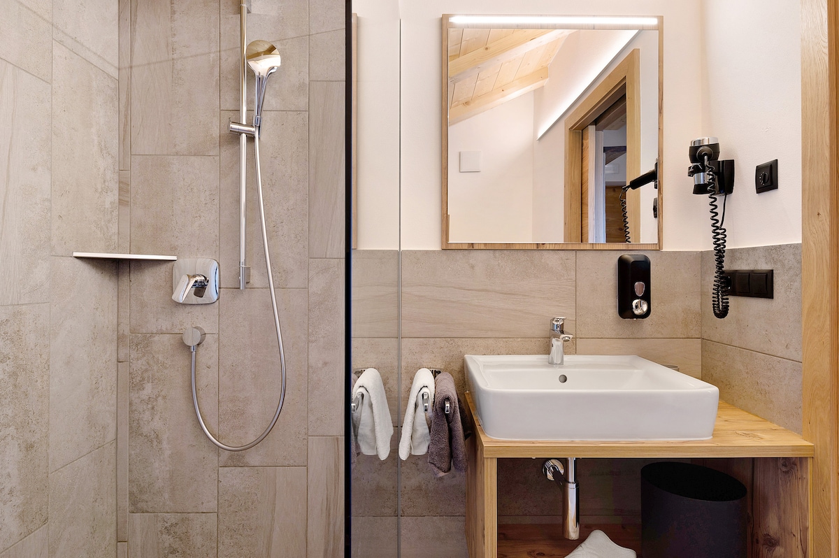 The bathroom features neutral-toned tiles and modern fixtures. A walk-in shower is equipped with a rainfall showerhead. A square sink is mounted on a wooden vanity, accompanied by a mirror. Two white towels and one dark towel hang nearby for convenience.