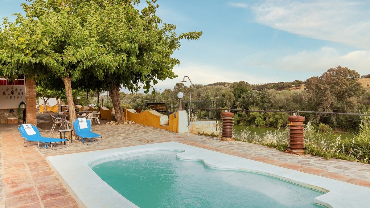 A private pool is set on a stone patio, surrounded by trees and greenery. A dining area with tables and chairs is visible in the background, along with two sun loungers positioned beside the pool, offering a serene view of the landscape.