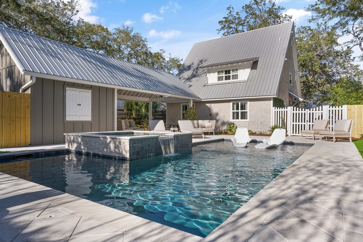 A spacious outdoor area features a shimmering pool with a connected hot tub. Lounge chairs are positioned around the pool, while a covered seating area and the home are visible in the background. Light reflects off the water, creating an inviting scene.