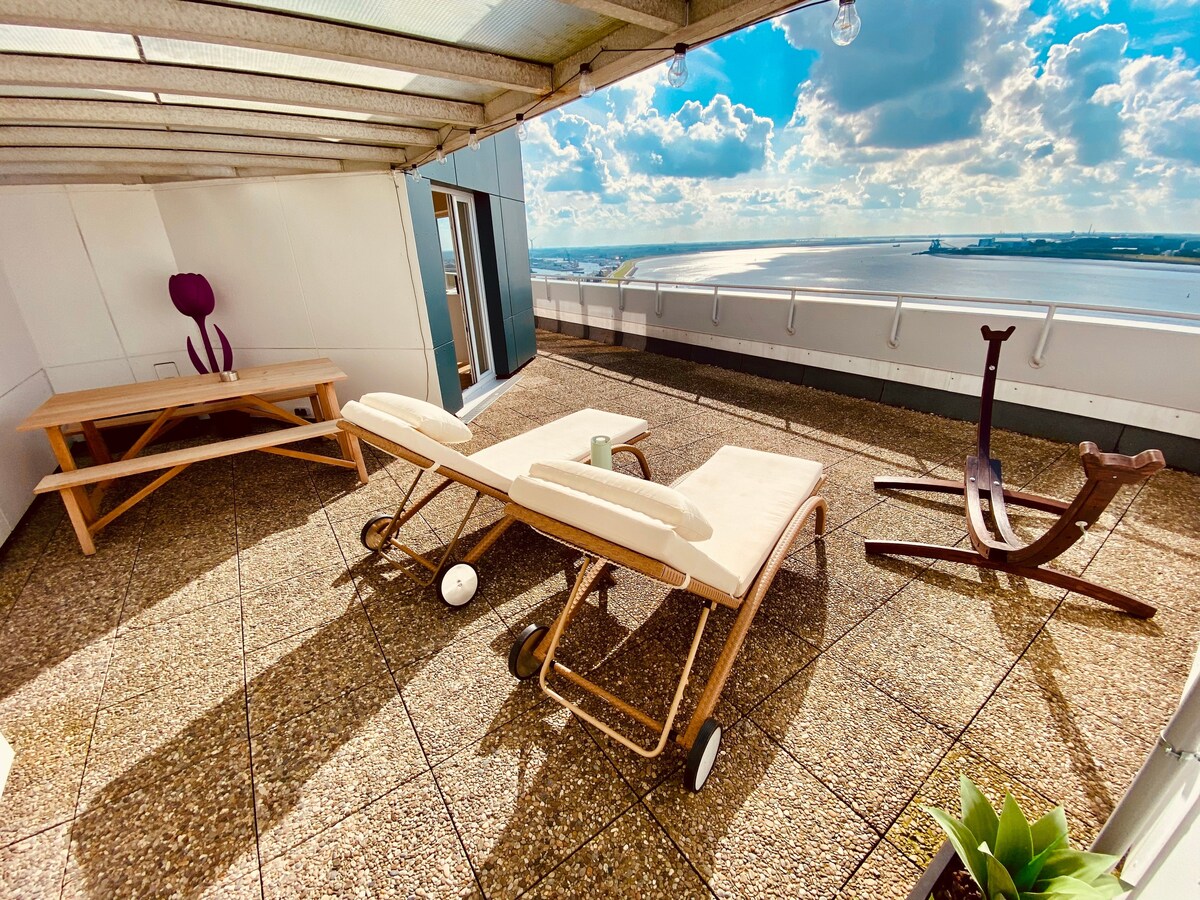 A private panorama terrace is featured with two relaxing loungers positioned for optimal views. A wooden dining table is positioned nearby, along with a decorative sculpture. The expansive space showcases a skyline view of the river and horizon under a bright, blue sky.