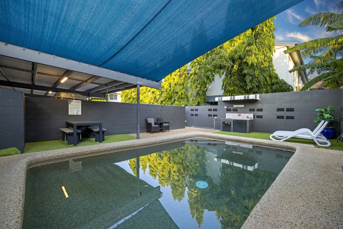 An outdoor pool area is surrounded by lush greenery and shaded by a blue canopy. A spacious seating area with a table and chairs is present, along with a barbecue station. The pool's clear water reflects the surrounding foliage, creating a calming atmosphere.