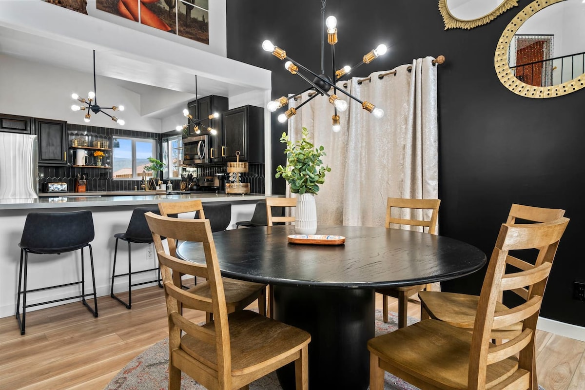 A round dining table with wooden chairs is positioned in an inviting space, accented by stylish lighting fixtures. The kitchen area features sleek black cabinetry and modern appliances. Natural light floods through a large window, enhancing the room's welcoming atmosphere.