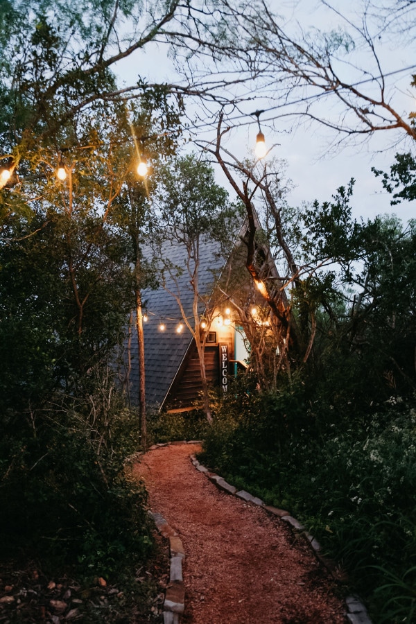 A winding gravel path leads to the entrance of a charming A-frame structure, surrounded by lush greenery and softly lit with string lights. The inviting facade features a wooden door, offering a serene atmosphere amid natural surroundings.