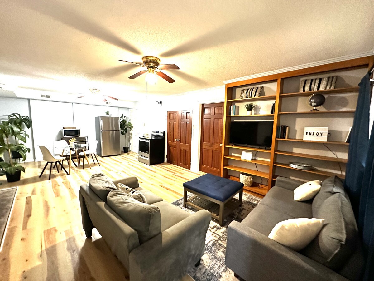 The living area features a cozy seating arrangement with two soft sofas and a central ottoman. A wooden shelving unit displays decorative items, while a large window allows natural light to brighten the space. A dining table for four is positioned beside a modern kitchen.