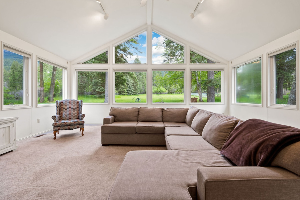 A spacious living area features a large sectional sofa, providing ample seating. The room is illuminated by natural light from tall windows, offering views of the surrounding greenery and yard. A single armchair complements the seating arrangement, creating an inviting space for relaxation.