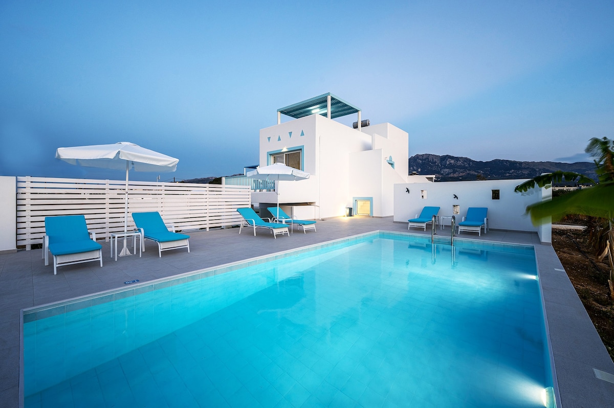 A serene swimming pool features crystal-clear water and is surrounded by blue lounge chairs. An umbrella provides shade nearby, while a modern villa with large windows stands in the background against a dusky sky.