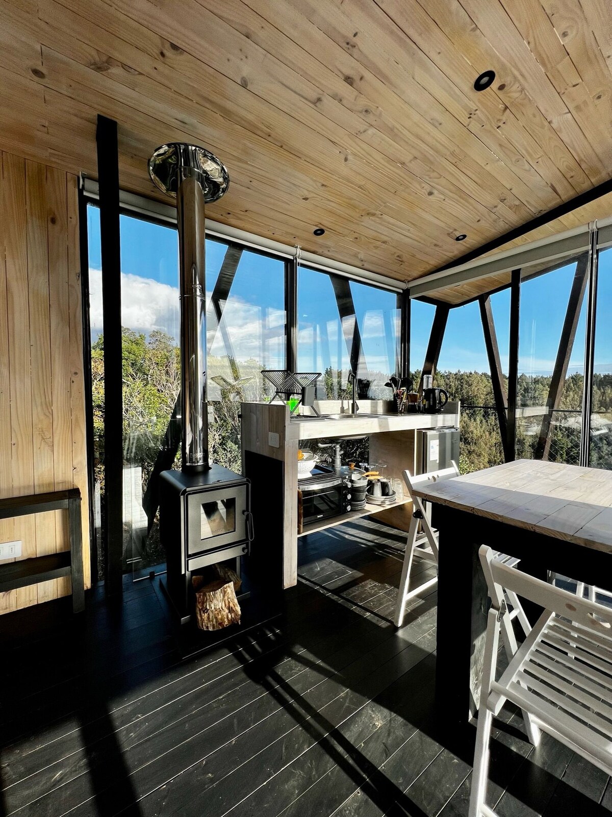 A modern kitchenette is featured with large windows offering natural sunlight and views of the surrounding forest. A compact stove is installed near a dining table with several chairs. Wooden paneling adorns the walls and ceiling, contributing to a cozy atmosphere.