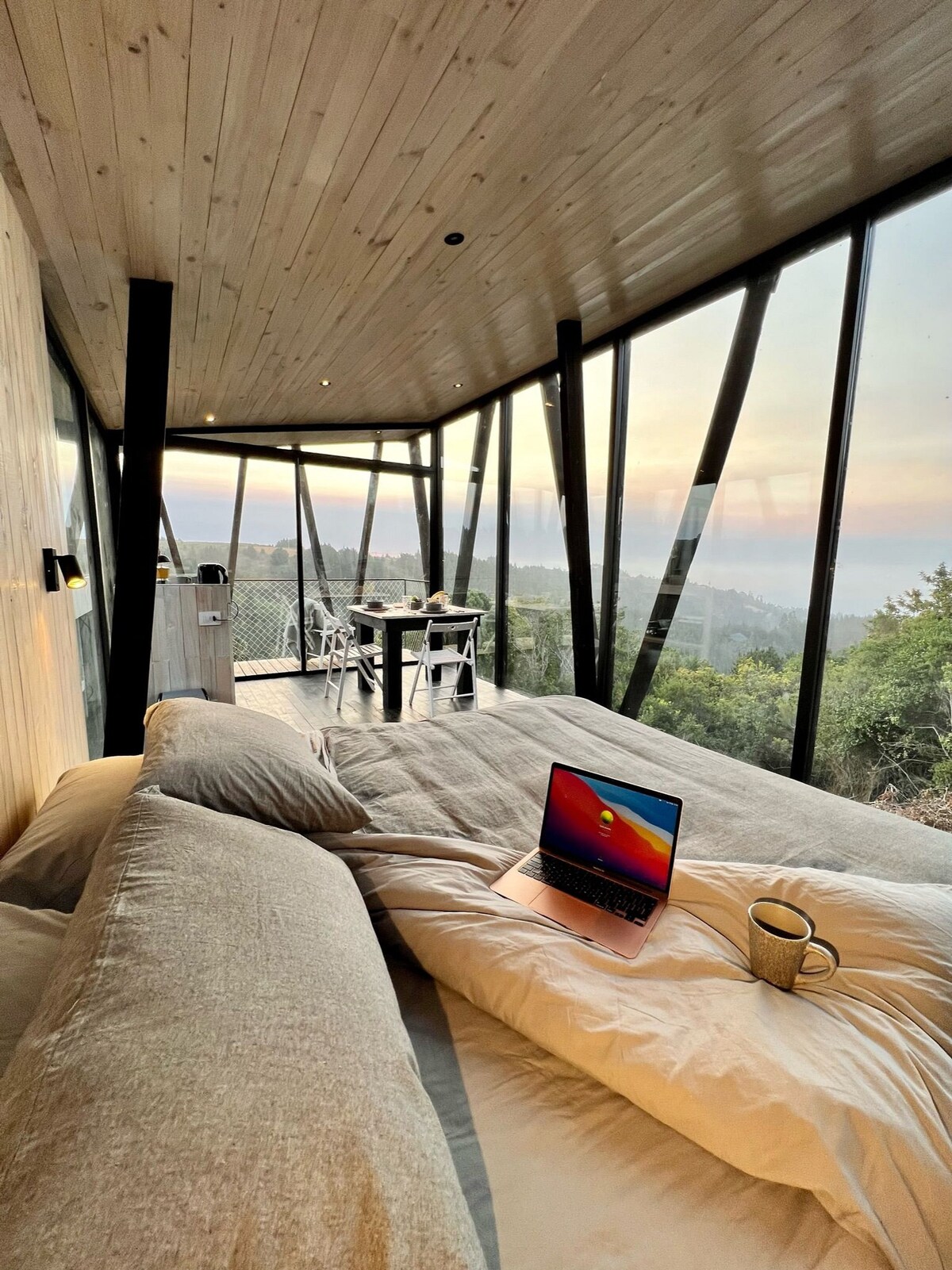 A cozy sleeping area is illuminated by soft natural light filtering through large glass walls. A spacious bed is adorned with neutral-toned linens. A laptop and a mug are placed on the bed, while a dining table with chairs is set against the backdrop of lush greenery.