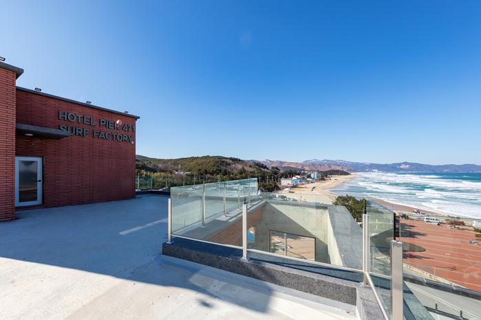 A rooftop area is visible with a clear view of the ocean and coastline. The surrounding landscape features sandy beach and rolling waves, while a brick building is positioned on one side. The space is unobstructed, offering an inviting outdoor atmosphere.