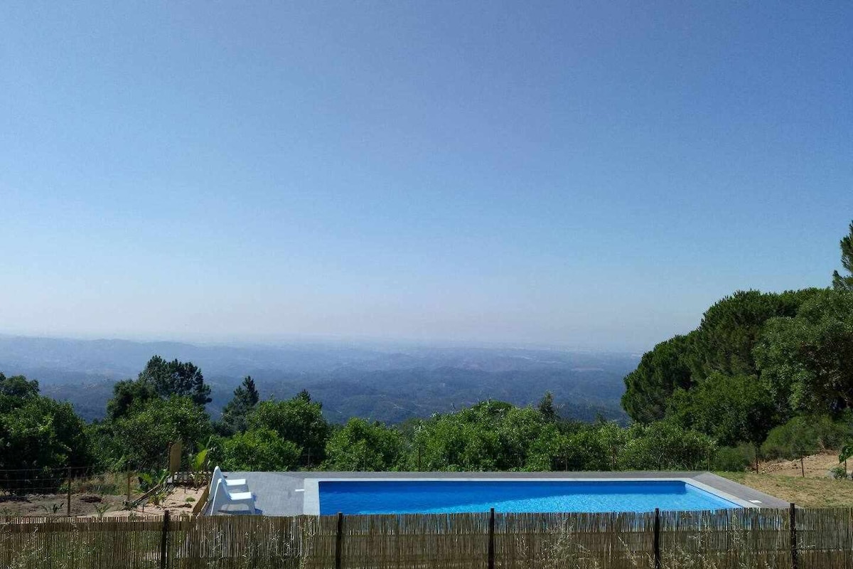A tranquil scene showcases a salt-water pool, framed by lush greenery. The panoramic view of rolling hills and valleys extends to the horizon, with a clear blue sky above. The serene environment invites relaxation and connection with nature.