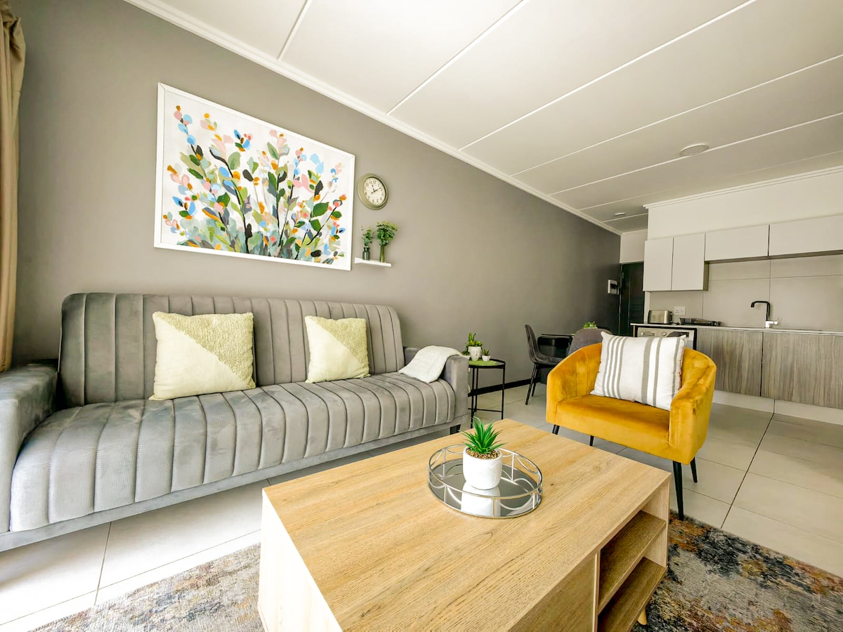 A modern living area showcases a grey upholstered sofa adorned with decorative cushions and a vibrant yellow accent chair. A wooden coffee table is centrally positioned, with a small potted plant on top. A colorful wall art piece adds a splash of color, while a clock and minimalist shelf enhance the decor.
